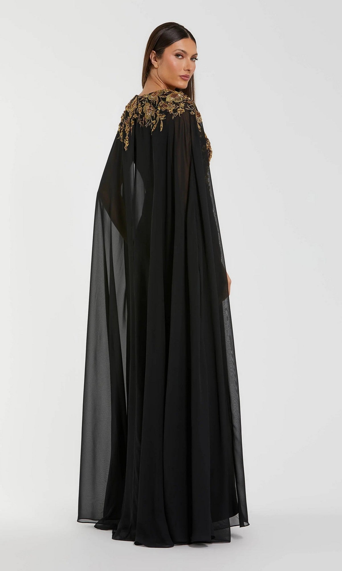 A woman stands facing backward in front of a plain white background, wearing the Long Formal Dress: Mac Duggal 13206—a black sheer gown with gold embroidery on the shoulders.